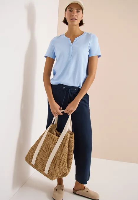 Frau im hellblauen Shirt und dunkelblauen Hosen mit großer Korbtasche und beigen Slippern vor hellem Hintergrund.