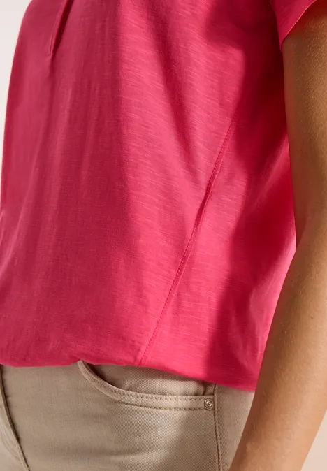 Close-up einer Person in leuchtend pinkem Oberteil und beigen Hosen, nur Torso und Arm sichtbar.