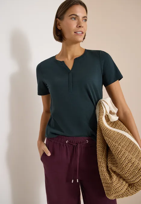 Basic Shirt im Tunika-Look deep petrol blue Frau in dunklem T-Shirt und weinroter Hose, trägt eine große, geflochtene Strohtasche.