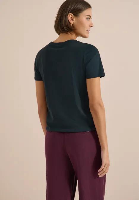 Basic Shirt im Tunika-Look deep petrol blue Person mit schulterlangem Haar, dunklem T-Shirt und lila Hose, stehend vor beigem Hintergrund.