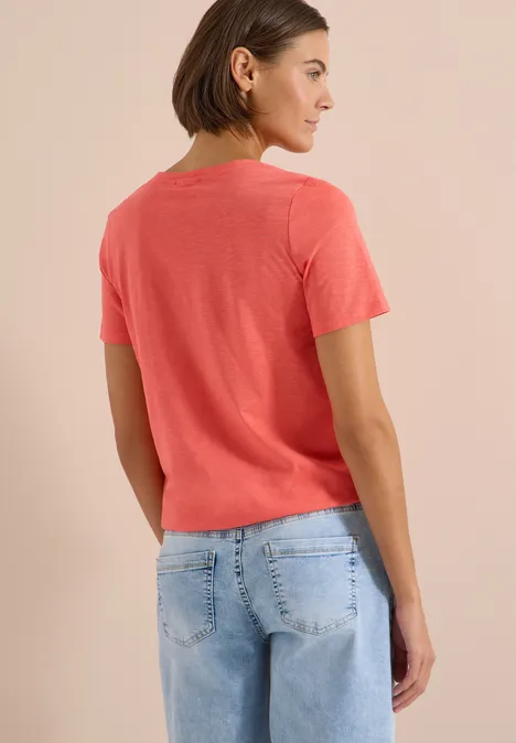 Basic Shirt im Tunika-Look shiny coral Frau steht von hinten, trägt ein korallenrotes T-Shirt und blaue Jeans vor neutralem Hintergrund.