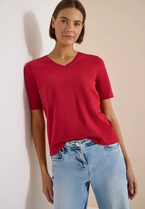 Eine Person in rotem T-Shirt mit V-Ausschnitt und hellen Jeans lehnt vor einer hellen Wand.