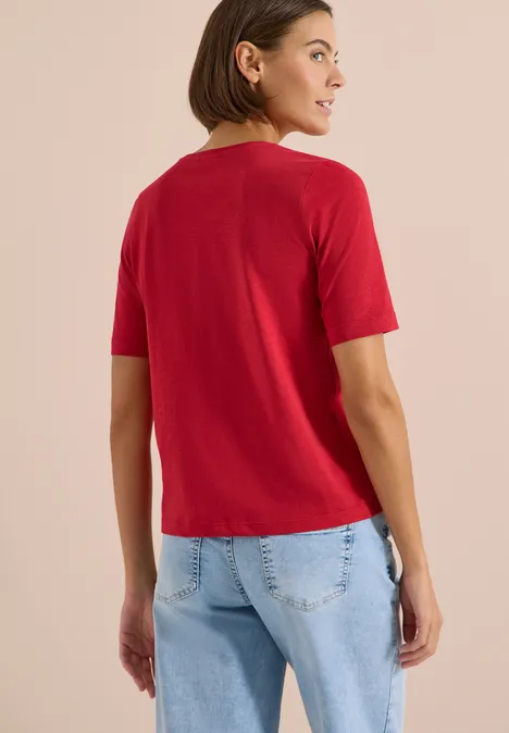 Person von hinten mit rotem T-Shirt und hellblauen Jeans vor neutralem Hintergrund.