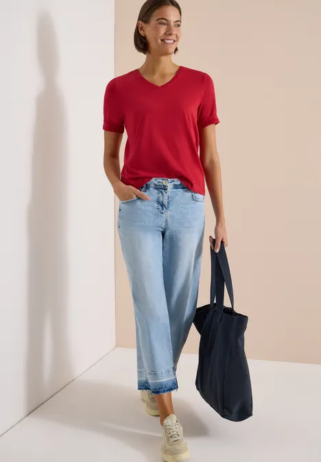 Eine Person mit rotem T-Shirt, hellblauen Jeans, weißen Sneakern und dunkelblauer Tasche vor hellem Hintergrund.