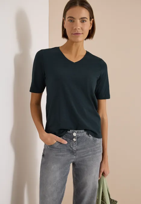 T-shirt basique à col V deep petrol blue Frau trägt dunkles T-Shirt und graue Jeans, hält grünes Tuch in der Hand vor weißem Hintergrund.
