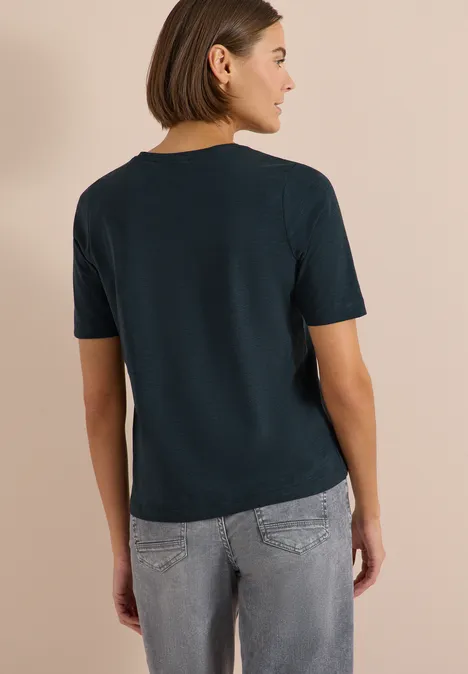 T-shirt basique à col V deep petrol blue Frau von hinten in marineblauem T-Shirt und grauer Jeans vor beigem Hintergrund.