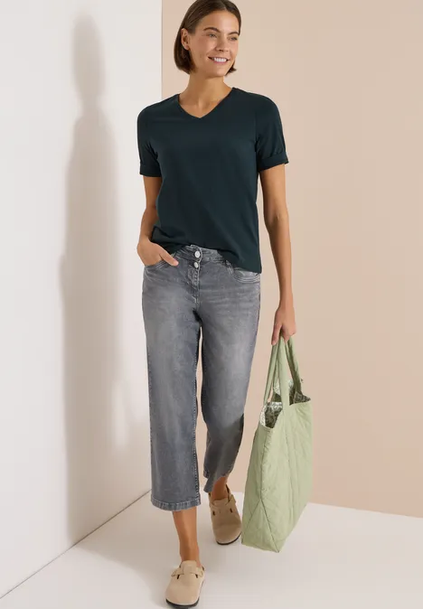 T-shirt basique à col V deep petrol blue Frau in schwarzem T-Shirt, weiten Jeans-Culottes und hellen Schuhen, trägt eine grüne Tragetasche.