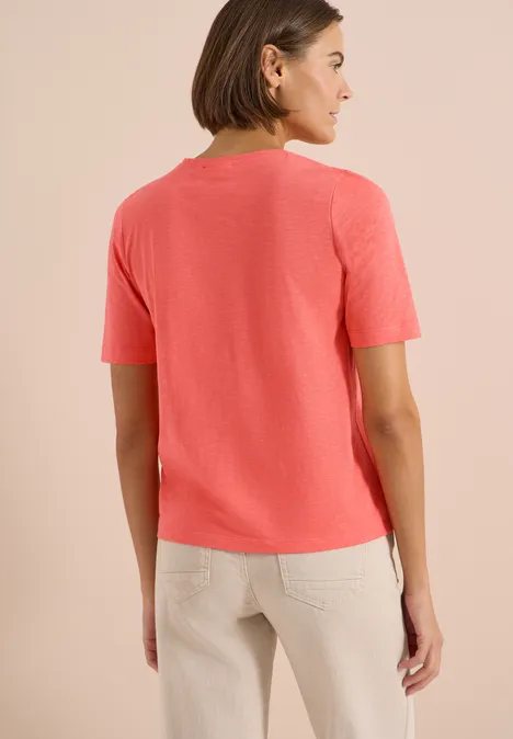 Frau von hinten in korallfarbenem T-Shirt und beige Hose vor hellem Hintergrund