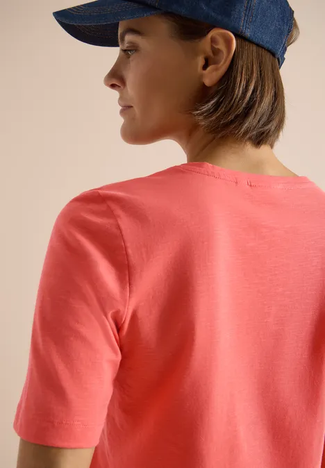 Rückenansicht einer Person mit korallfarbenem T-Shirt und blauer Kappe vor beigem Hintergrund.