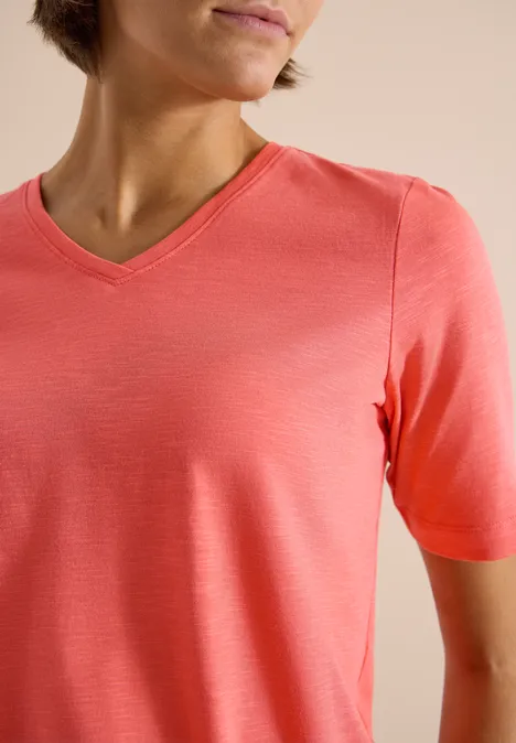 Person trägt ein korallenfarbenes V-Ausschnitt-Shirt vor neutralem Hintergrund.