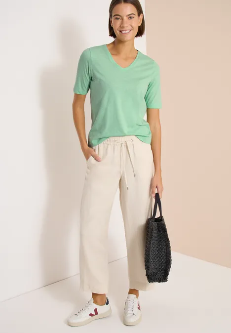 Person steht vor Wand, trägt hellgrünes V-Shirt, helle weite Hose, weiße Sneaker, hält schwarze Tasche.