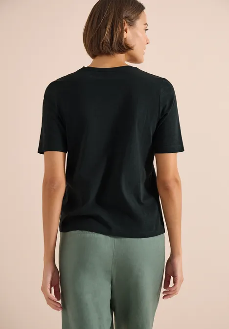 Frau von hinten in schwarzem T-Shirt und grünen Hosen vor neutralem Hintergrund