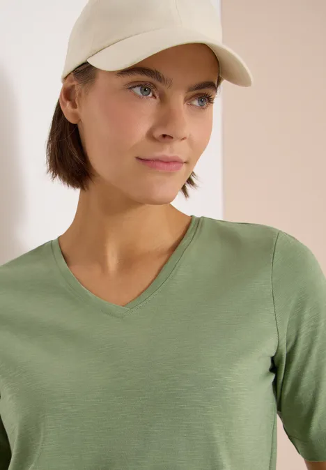 Frau in olivgrünem V-Shirt und hellbeiger Kappe vor neutralem Hintergrund.