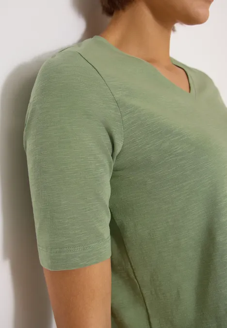 Frau in hellgrünem V-Ausschnittshirt mit kurzen Ärmeln, seitliche Ansicht, oberer Oberkörper sichtbar.