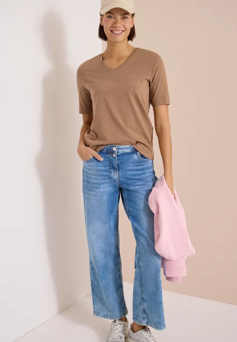 Basic T-Shirt mit Kurzarm und V-Neck creamy mocha Frau in braunem T-Shirt und blauer Jeans hält rosa Pullover vor hellem Hintergrund.