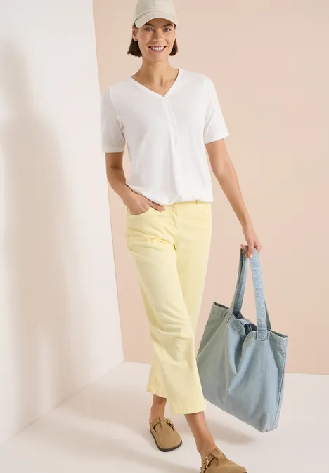 Vrouw in wit V-T-shirt, gele korte broek, loafers en spijkertas voor een lichte, minimalistische achtergrond.
