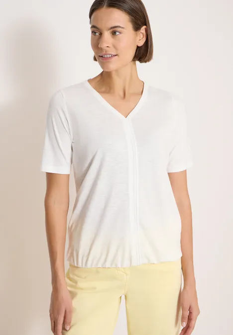 Een vrouw draagt een wit V-hals T-shirt en een gele broek tegen een effen achtergrond.