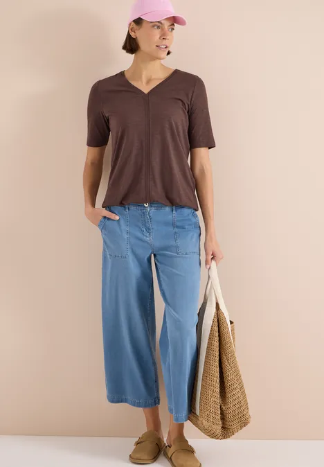 Kurzarm T-Shirt im Tunika-Look brown cacao Frau mit braunem V-Shirt, weiten blauen Culotte-Hosen und Strohtasche.