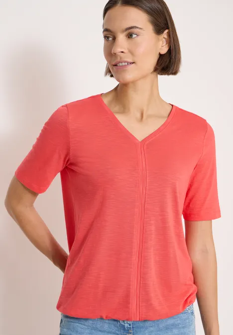 Une personne au visage flou porte un t-shirt corail à col en V et manches courtes.