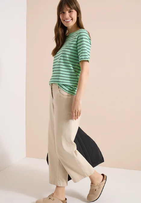 Frau im Profil trägt gestreiftes grünes Oberteil, helle weite Hosen und Slip-ons vor neutralem Hintergrund.