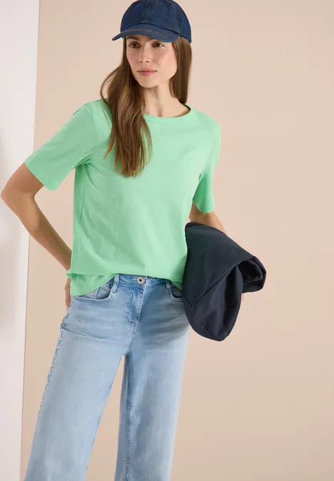 Eine Person mit grünem T-Shirt, Jeans und Basecap hält eine Jacke in der Hand.