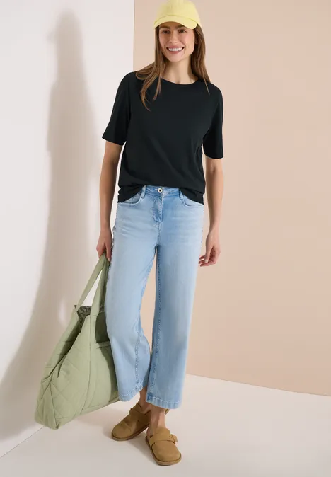 Frau in schwarzem T-Shirt, geraden blauen Jeans, braunen Slippern, trägt große hellgrüne Tasche.