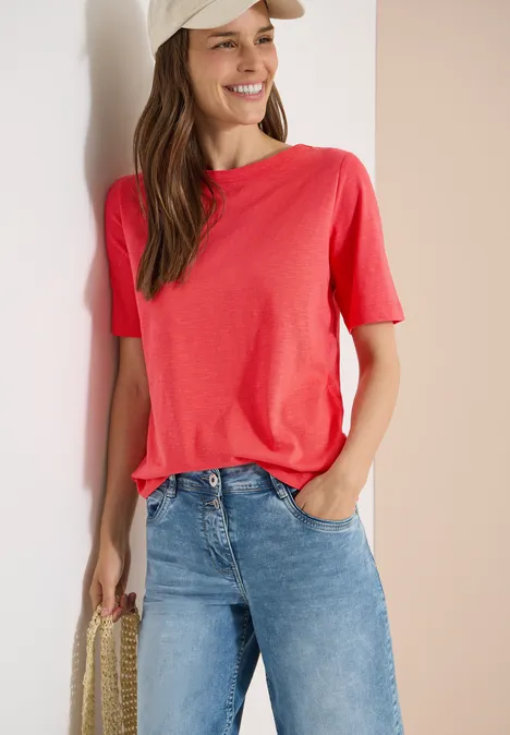 Frau lehnt an weißer Wand, trägt rosa T-Shirt, blaue Jeans und hält eine geflochtene Tasche.