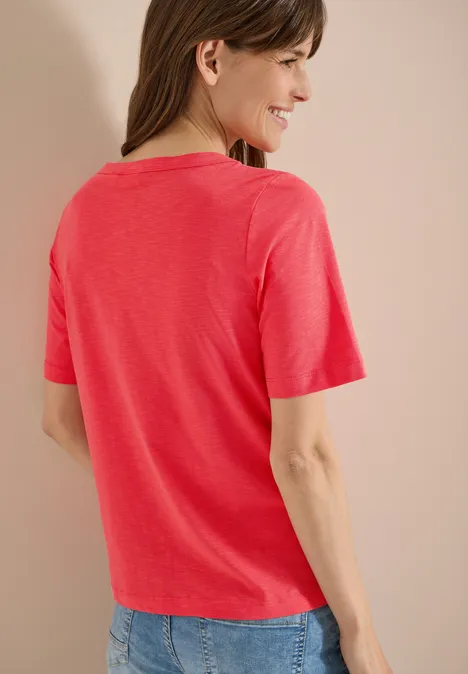 Rückansicht einer Frau in korallrotem T-Shirt und blauer Jeans vor neutralem Hintergrund.