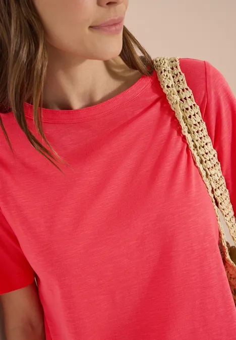 Nahaufnahme einer Frau in korallfarbenem T-Shirt mit gewebtem Strohtaschen-Träger.