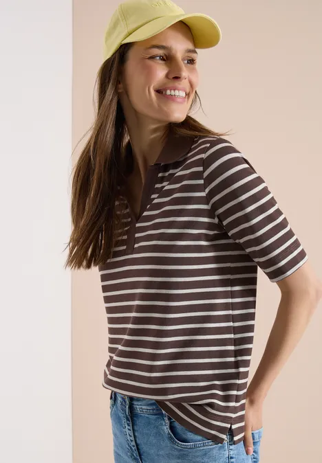 Eine Person trägt eine gelbe Cap, ein gestreiftes braun-weißes T-Shirt und Jeans vor neutralem Hintergrund.