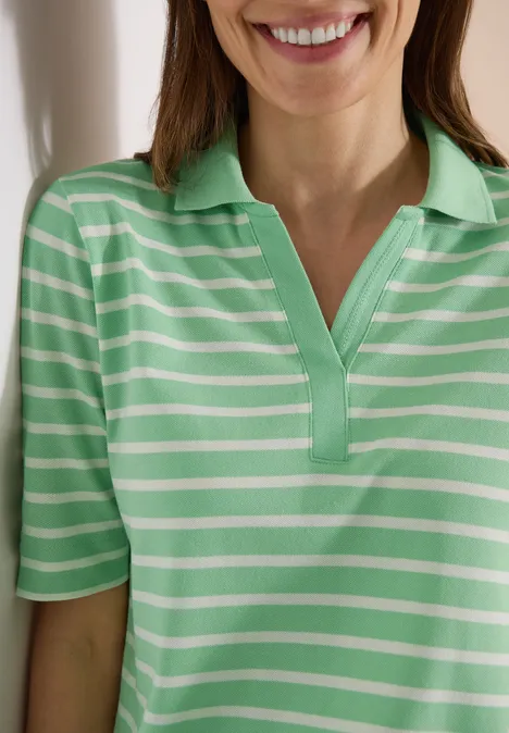 Eine Frau lehnt lächelnd an einer Wand und trägt ein grün-weiß gestreiftes Poloshirt.