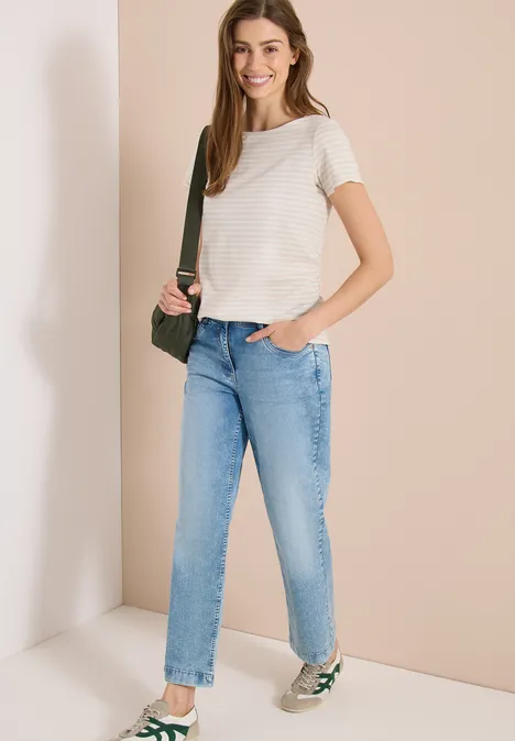 Junge Frau trägt weiß gestreiftes T-Shirt, blaue Jeans, grüne Umhängetasche und Sneaker.