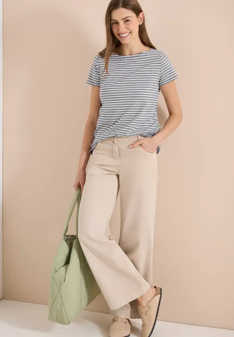 Frau in gestreiftem T-Shirt, weiten beigen Hosen, grüner Tasche und beigen Slippern vor neutralem Hintergrund