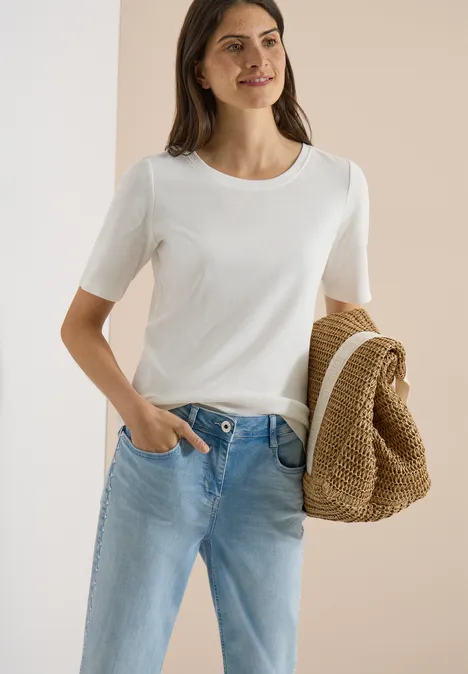 Basic geribd Shirt vanilla white Vrouw in wit T-shirt en lichtblauwe jeans houdt een gevlochten strohoed vast voor een neutrale achtergrond.