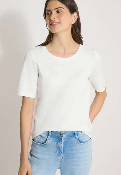Basic geribd Shirt vanilla white Vrouw draagt wit T-shirt en lichtblauwe jeans, hand in de zak, neutrale achtergrond.