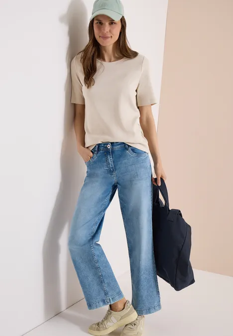 Frau in weißem T-Shirt und weitem Jeans, trägt Mütze und Tasche, lehnt elegant an heller Wand.