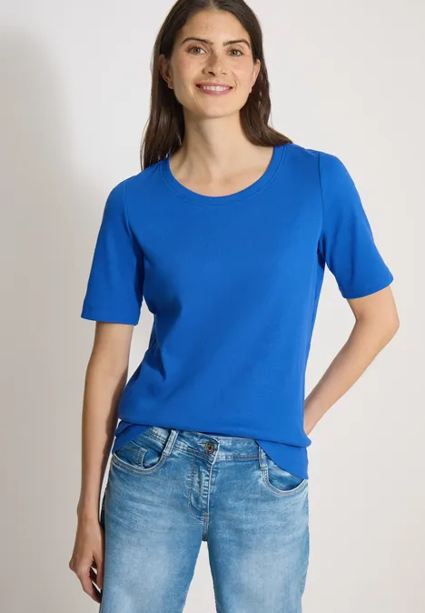 Basic Shirt met geribde textuur surf blue Vrouw in een koningsblauw T-shirt met korte mouwen en lichtblauwe jeans tegen een neutrale achtergrond.