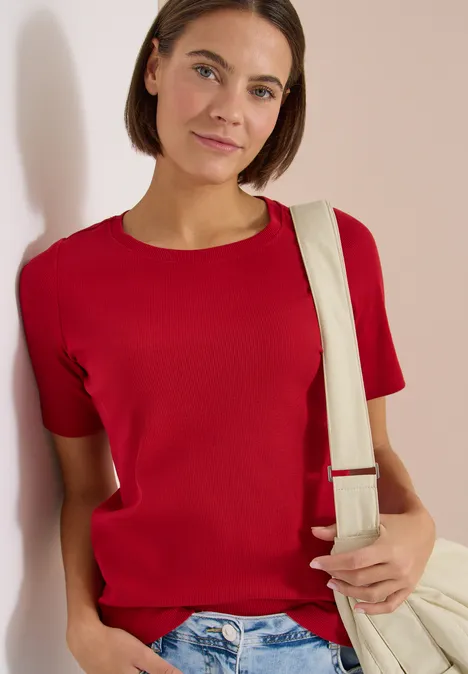 Basic T-Shirt in Rippstruktur tamarillo red Person lehnt an Wand, trägt rotes T-Shirt, blaue Jeans und eine helle Schultertasche.
