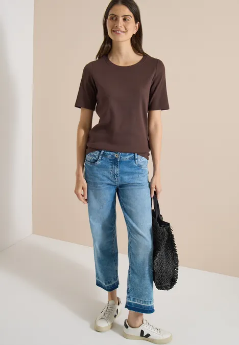 Basic Shirt met geribde textuur brown cacao Vrouw in bruin T-shirt en blauwe jeans, witte sneakers, zwarte tas, staand voor een lichte achtergrond.