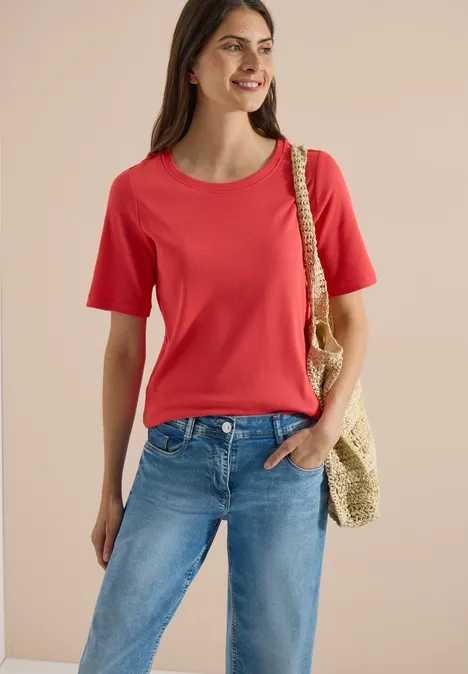 Basic Shirt met geribde textuur coral Vrouw in rood t-shirt en blauwe spijkerbroek met strooien tas over de schouder, glimlachend.