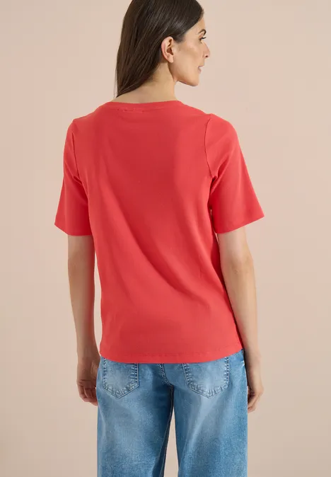Basic Shirt met geribde textuur coral Een persoon van achteren in een rood T-shirt en blauwe spijkerbroek tegen een neutrale achtergrond.