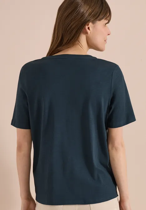 Frau in dunkelgrünem kurzärmeligem Oberteil, Rückenansicht vor neutralem Hintergrund.