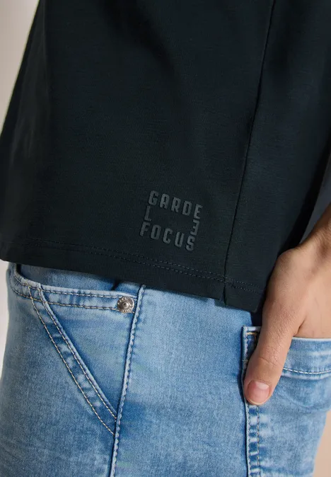 Schwarzes Shirt mit Aufdruck "GARDE FOCUS" und blauem Jeans, Hand in der Hosentasche.