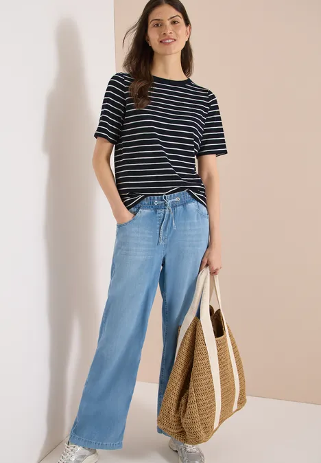 Een persoon draagt een gestreept T-shirt, wijde jeans en houdt een gevlochten tas vast.