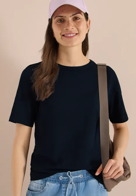 Vrouw in donkerblauw T-shirt met korte mouwen, bruine schoudertas, effen beige achtergrond.