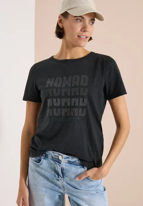 Kurzarm Shirt im Ausbrenner-Look palm breeze green Frau mit unkenntlichem Gesicht trägt dunkelgraues T-Shirt mit 'NOMAD'-Print und blaue Jeans vor hellem Hintergrund