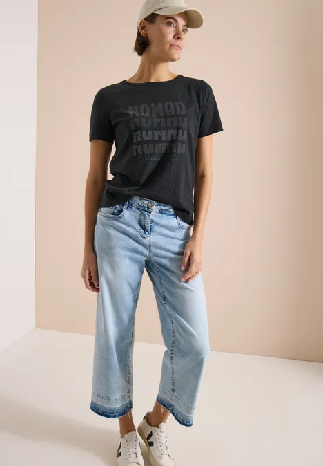 Kurzarm Shirt im Ausbrenner-Look palm breeze green Modisches Outfit: Person trägt schwarzes T-Shirt mit Aufdruck, helle weite Jeans, weiße Sneaker und Cap.