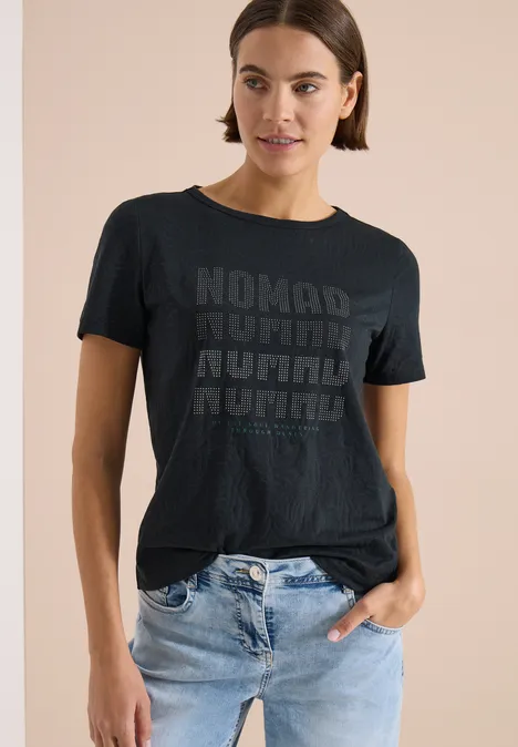 Kurzarm Shirt im Ausbrenner-Look palm breeze green Eine Person trägt ein schwarzes T-Shirt mit wiederholtem 'NOMAD'-Schriftzug und helle Jeans.