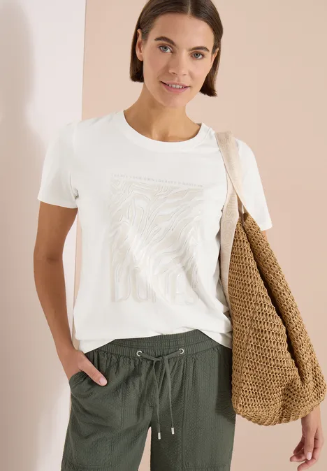 Frau in weißem T-Shirt, grüner Hose, großer Korbtasche über der Schulter, vor hellem Hintergrund.