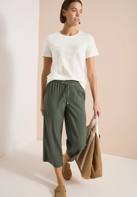 Frau in weißem T-Shirt, weiten, grünen 3/4-Hosen, Basttasche und hellbraunen Slippern vor hellem Hintergrund.